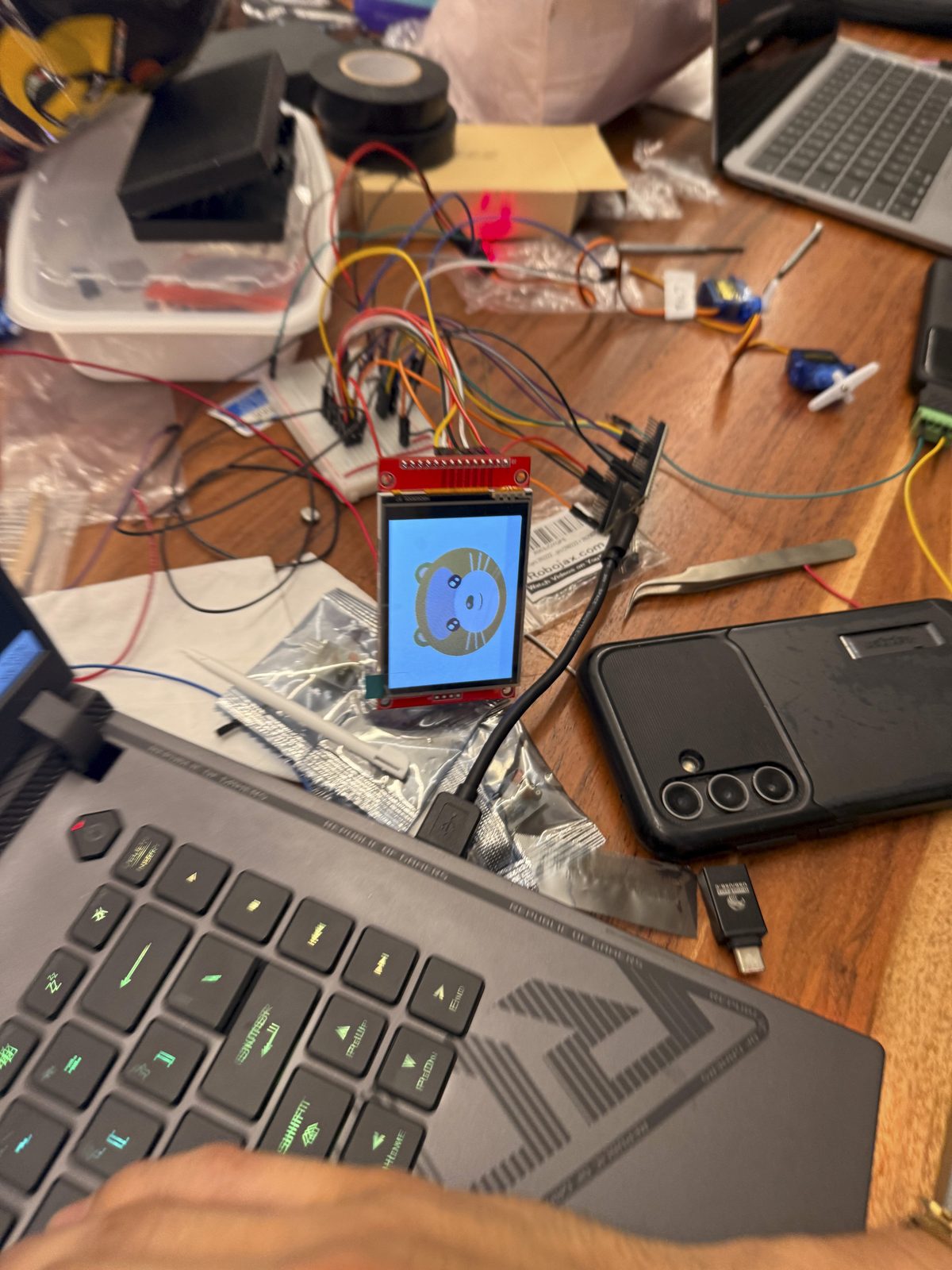 The screen wired on a breadboard, showing the otter in inverted colors, with servos, a laser and a laptop around it.