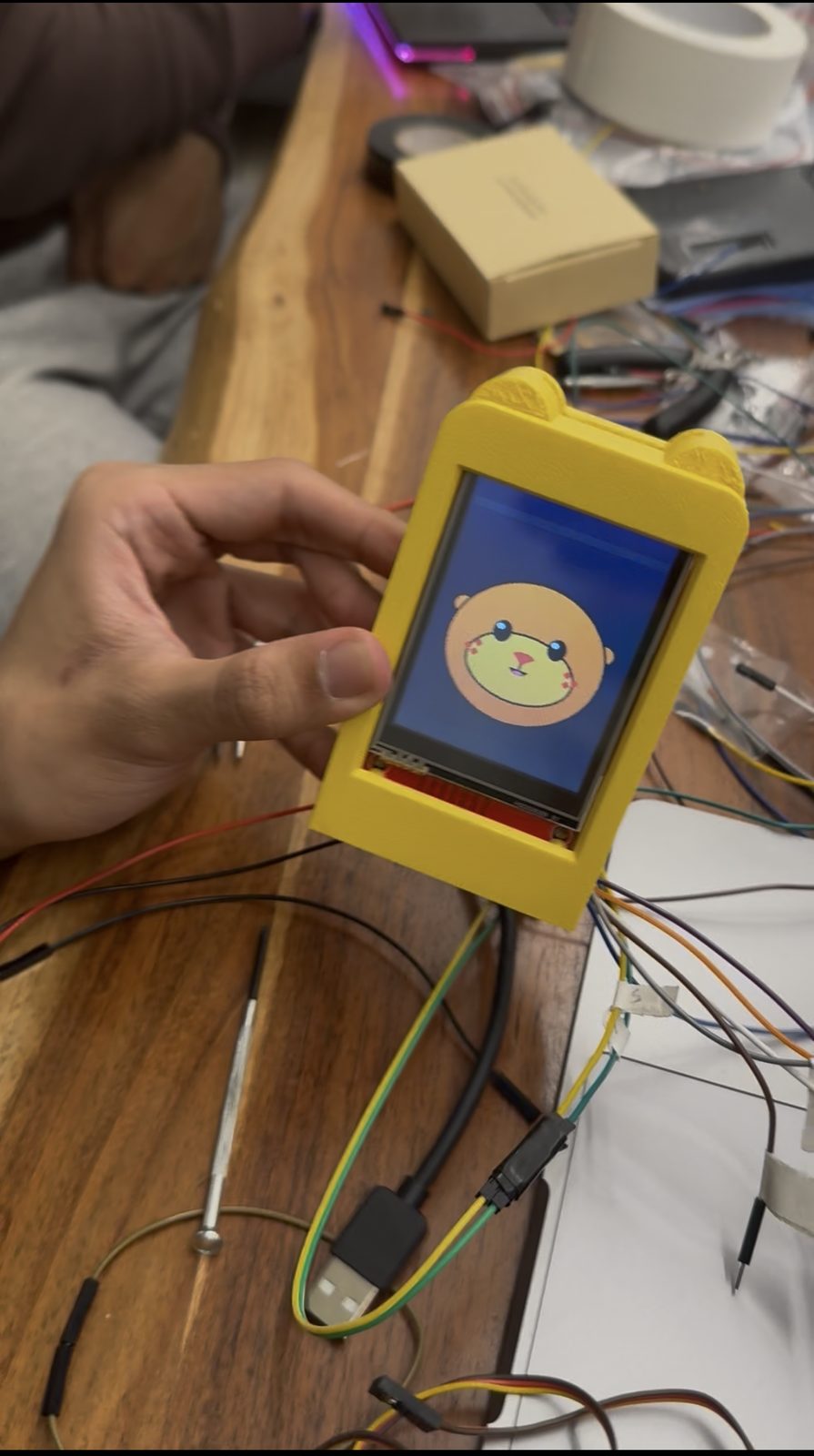 A hand holding the yellow 3D-printed case with otter ears, the screen inside showing the golden otter.