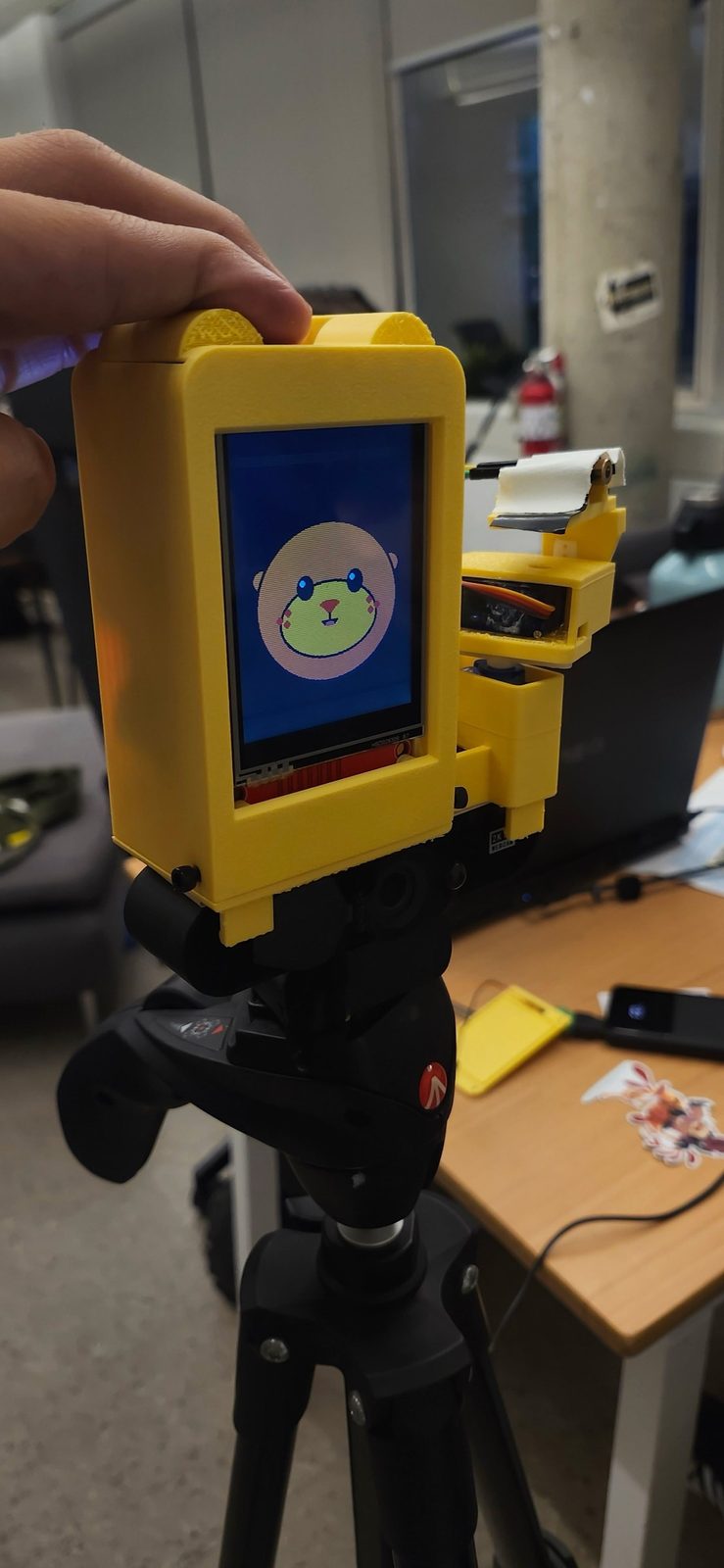 The finished yellow Otter Tutor robot seen from the front, clipped onto a webcam on a tripod, with the otter's face on its screen and the laser arm on the right.