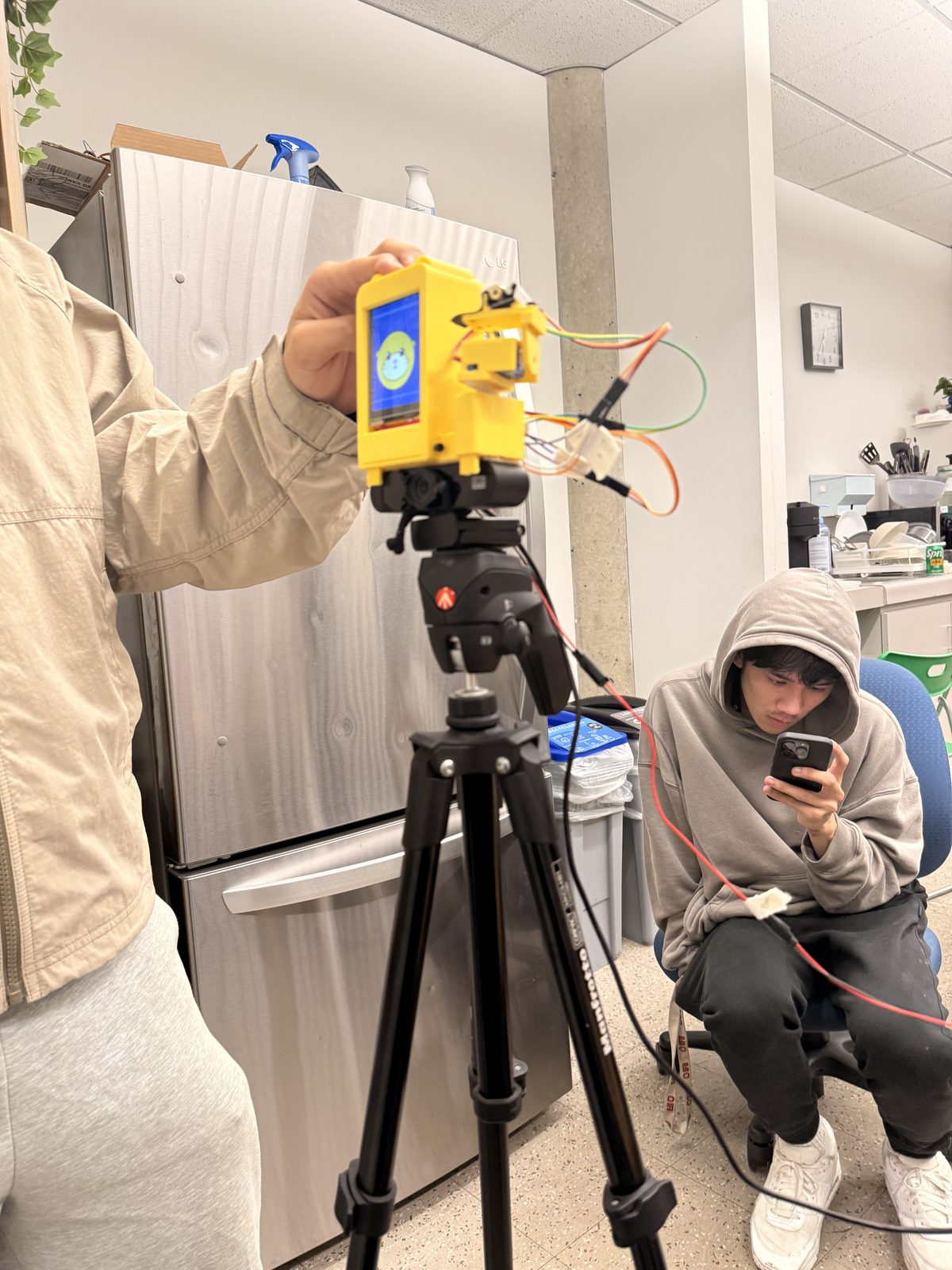 The finished robot clipped onto a webcam on a tripod, held up for a test while a teammate checks it from his phone.